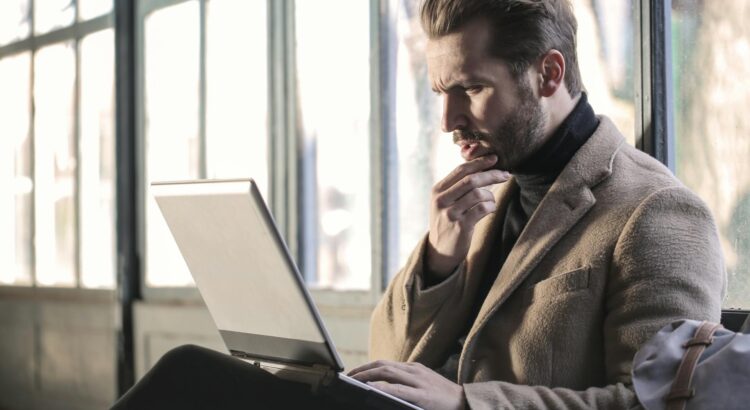 Mann sitzt vor einer Fensterfront und schaut in einen geöffneten Laptop